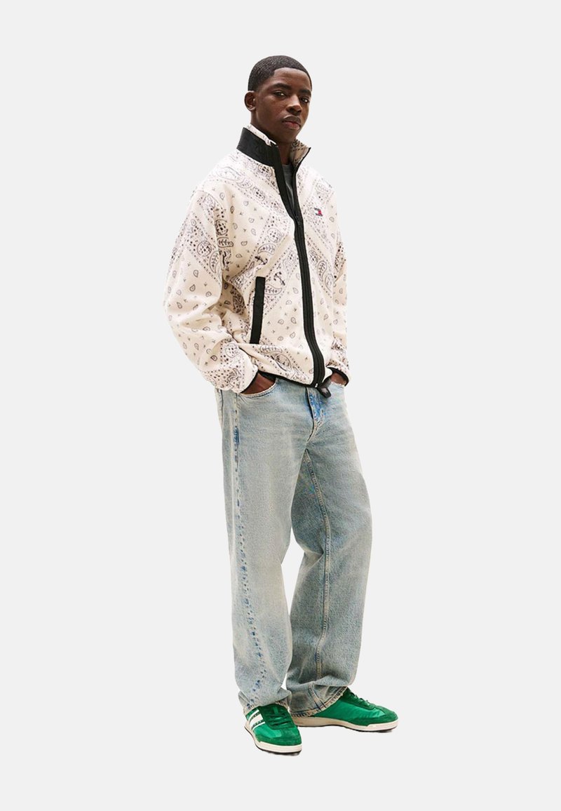 Chaqueta blanca con estampado y cremallera con detalles en negro, jeans de mezclilla azul claro y zapatillas verdes. La chaqueta presenta diseños intrincados en una tela de peso medio.
