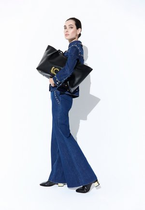 Mujer con chaqueta vaquera azul con tachuelas y vaqueros acampanados que sostiene un gran bolso negro de cuero con logo dorado, y lleva botas negras con tacones dorados.