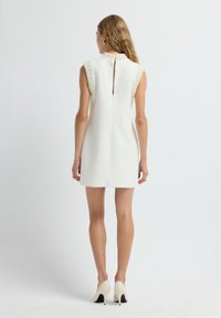 Vrouw die met haar rug naar de camera staat in een mouwloze witte jurk met pareldetails en witte hoge hakken, tegen een effen achtergrond.