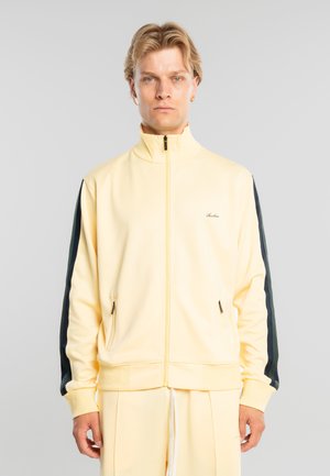 Mand med kort blondt hår iført en bleg gul lynlåstrøje med sorte striber på ærmerne og matchende bukser, stående neutralt.