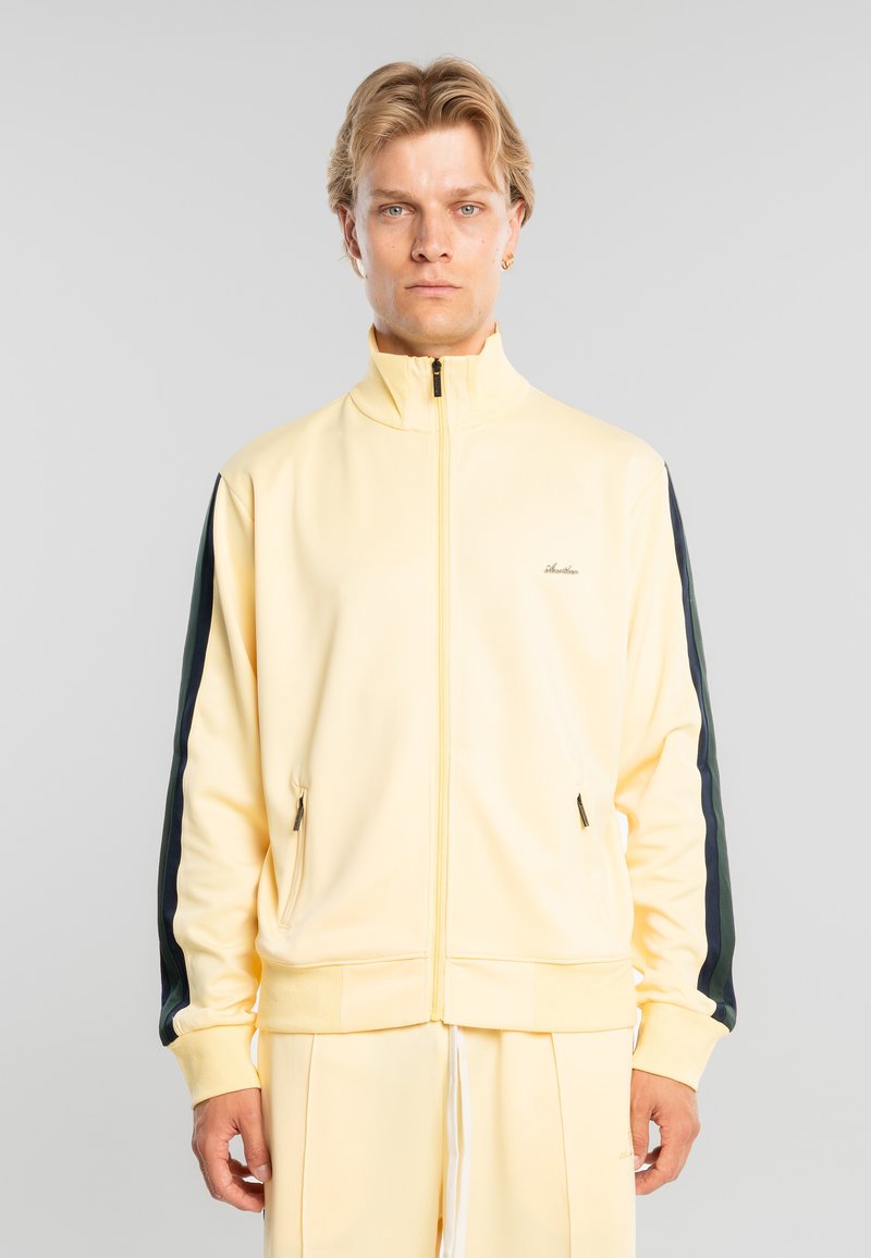 Hombre con cabello rubio corto que lleva una chaqueta pale amarilla con cremallera y rayas negras en las mangas, y pantalones a juego, está de pie en posición neutral.