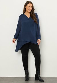Top blu navy a maniche lunghe con scollo a V e orlo asimmetrico, realizzato in tessuto leggero. Indossato con pantaloni aderenti neri e stivali neri con tacco.