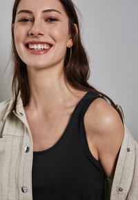 Top nero senza maniche con scollatura rotonda, abbinato a una leggera camicia a righe. Il modello ha capelli lunghi e mossi e grandi orecchini a cerchio.