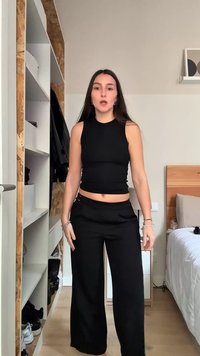 Nuori nainen, jolla on pitkät tummat hiukset, seisoo makuuhuoneessa päällään musta hihaton lyhyt paita ja väljät mustat housut.