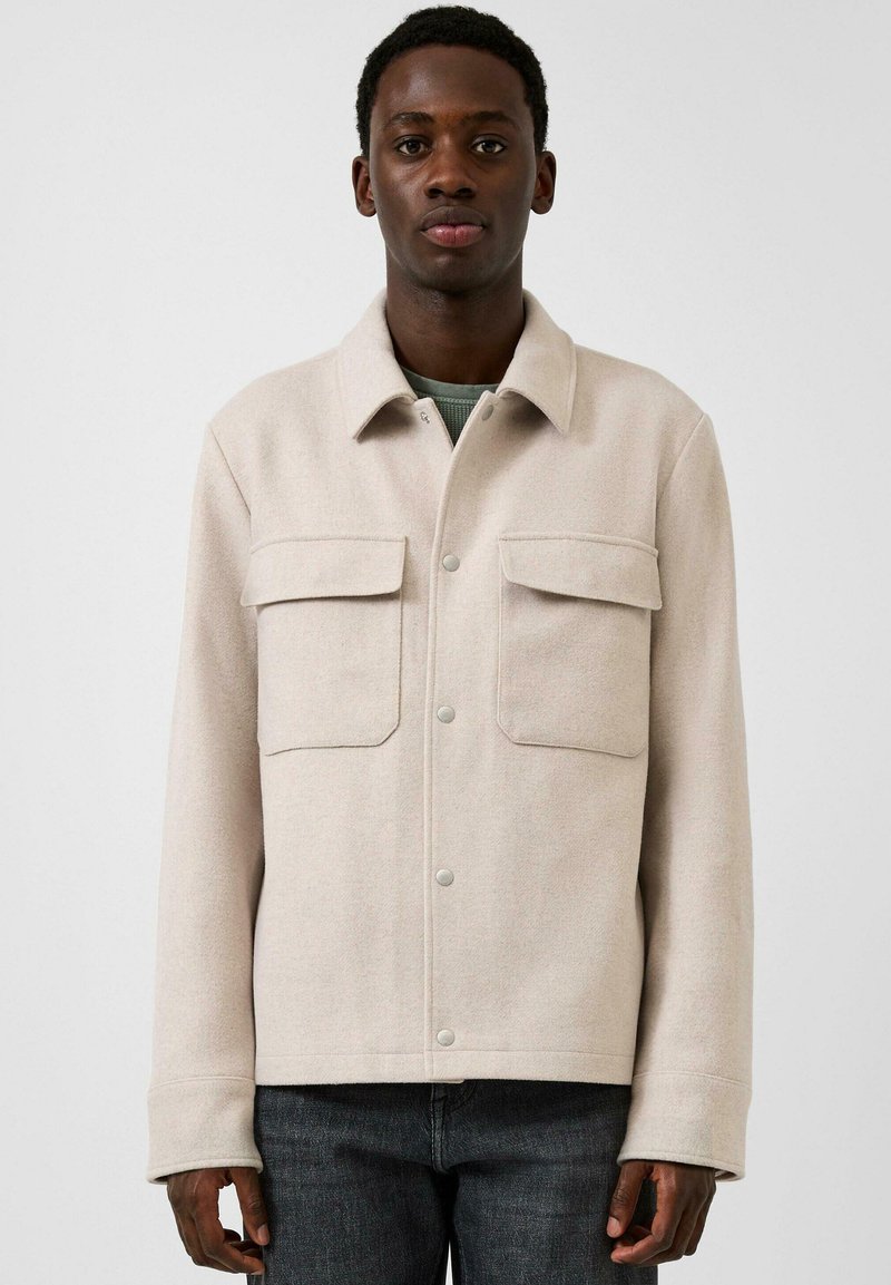 Man met een crème-knoopjasje met grote klepzakken, staand tegen een effen lichtgrijze achtergrond.