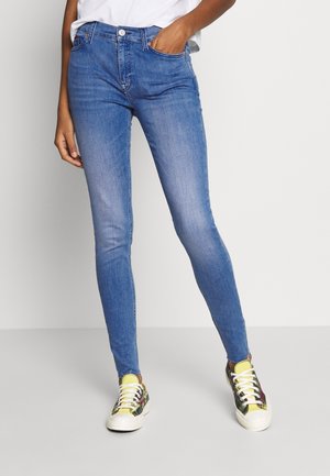 Personne portant un jean bleu slim et des baskets multicolores avec des lacets blancs, debout avec une main dans la poche devant un fond uni.