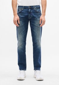 Jeans in denim blu scuro con un design a gamba dritta, dettagli sbiaditi sulle cosce, cinque tasche e cuciture in contrasto. Indossati con scarpe da ginnastica bianche.