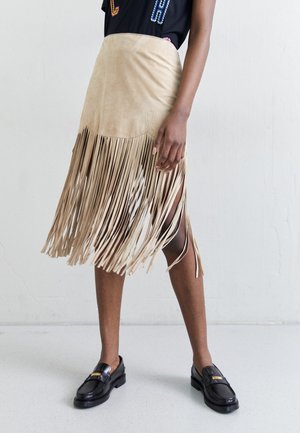 Beige nubukseelik, mille allääres on pikad frangid, sujuva tekstuuriga ja külgmiste lõigetega, koos mustade loafers'itega, millel on kuldne aktsent.