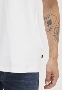 Witte katoenen t-shirt met korte mouwen, zichtbare stiksels en een zijlabel, gecombineerd met donkerblauwe spijkerbroek en een tatoeage op de arm.
