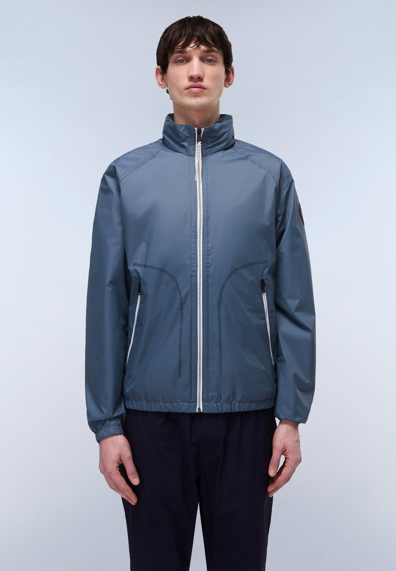 Lichte, blauwe windjack met een hoge kraag, voorritssluiting en elastische mouwen. Voert twee zijzakken met zwarte accenten.
