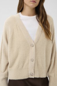 Žena nosící béžový chlupatý cardigan se třemi ozdobnými knoflíky přes bílé žebrované tričko a tmavě hnědou sukni nebo kalhoty.