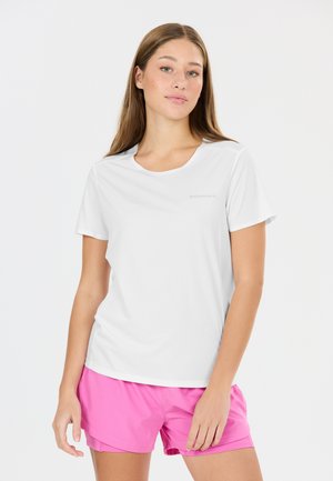 Ung kvinde med langt hår iført en hvid kortærmet sportstrøje og lyserøde sportsshorts, stående mod en ensfarvet baggrund.