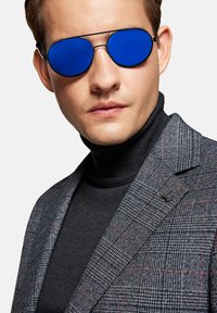 Okulary przeciwsłoneczne w niebieskiej tonacji z elegancką czarną metalową oprawką. Model nosi czarny golf i szary żakiet w kratę z czerwonymi akcentami.
