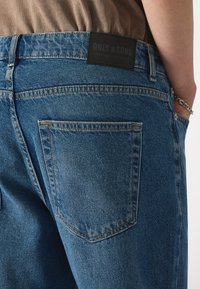 Jean en denim bleu avec une finition texturée, présentant une étiquette de marque en cuir noir, deux poches arrière et des coutures orange contrastées.