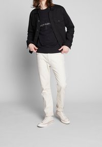 Černá denimová bunda, černý svetr s logem Calvin Klein, krémové kalhoty a bílé tenisky; uvolněný střih, jednoduchý design a minimální akcenty.