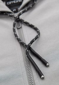 Veste zippée grise avec cordons noirs à extrémités argentées, fabriquée en tissu texturé avec un gros plan des détails de couture.