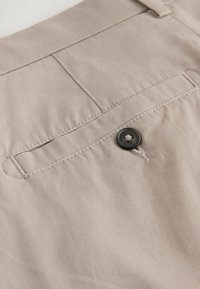 Nahaufnahme einer beigen Hosentasche mit genähten Details und einem einzigen dunklen Knopfverschluss.