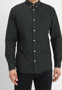 Muž oblečený v tmavě zelené a černé kostkované košili s dlouhým rukávem, s bílými knoflíky, v kombinaci s černými kalhotami.