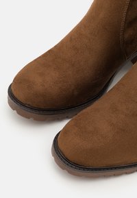 Bottines en daim marron avec bout arrondi, semelle en caoutchouc noire contrastante et coutures discrètes le long des bords. Finition texturée.