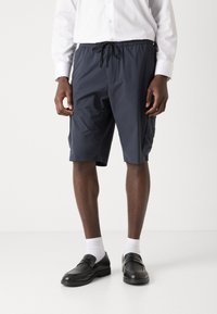 Shorts légers de couleur marine en matériau synthétique avec une taille élastique, cordon de serrage et poches latérales. Portés avec des mocassins noirs et des chaussettes blanches.
