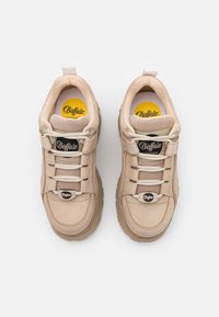 Beżowe sneakersy z masywną podeszwą, wykonane z gładkich i teksturowanych materiałów. Posiadają okrągłe sznurowadła oraz wyraźne czarne oznaczenia po bokach.