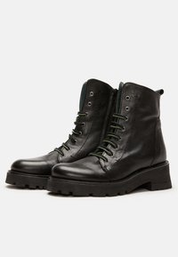 Botas de tobillo de cuero negro con acabado texturizado, cordones verdes y suela de goma gruesa. Presentan un cierre de cremallera en la parte delantera y un diseño de puntera redonda.