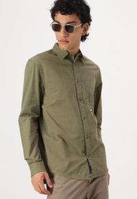 Olive green long-sleeve shirt with a button-down collar, front pocket, and embroidered logo. Smooth cotton fabric with a relaxed fit.