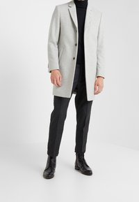 Abrigo gris de mezcla de lana con un ajuste entallado, solapas con muescas y cierre de un solo botón, combinado con pantalones oscuros y botas negras con cordones.