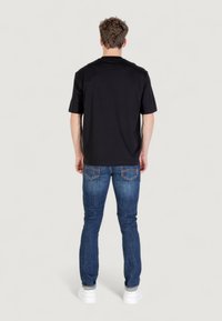 Hombre vestido con una camiseta negra de manga corta, jeans azules y zapatillas blancas, de pie dando la espalda contra un fondo claro y liso.