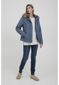 Marineblå quilted jakke med faux fur hætte, der har en lynlåslukning, sidelommer og en tekstureret cremefarvet sweater under, parret med blå jeans.