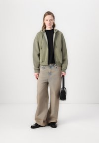 Olijf groene zip-up hoodie over een zwarte top, gecombineerd met lichtgewassen wijde spijkerbroek. Een zwarte handtas en schoenen maken de look compleet.