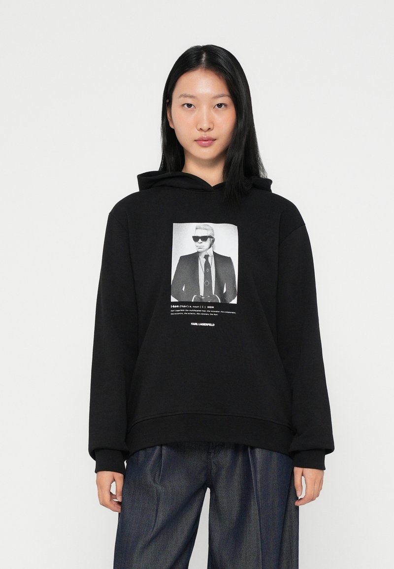 Fekete kapucnis pulóver puha anyagból, amelyen egy öltönyös férfi napszemüveggel látható, szöveggel kísérve, laza fazonban.