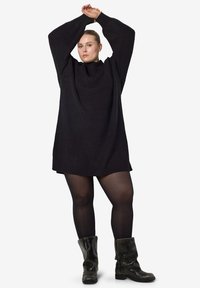 Μαύρο oversized πλεκτό φόρεμα με ψηλό λαιμό, μακριά μανίκια και ριμπ υφή, συνδυασμένο με γυαλιστερές μαύρες μπότες μέχρι το γόνατο.