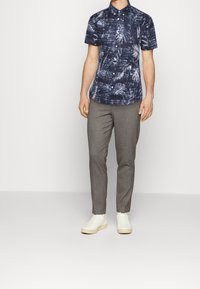 Chemise bleu marine à manches courtes avec un motif de feuilles blanches, assortie à un pantalon gris ajusté et des baskets blanches. Tissu lisse et coupe ajustée.