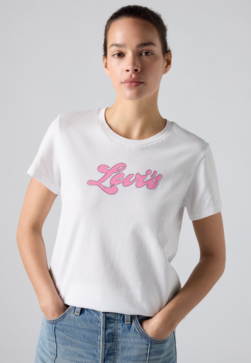 Femme portant un t-shirt Levi’s blanc avec un logo rose, debout les mains dans les poches d’un jean bleu, sur un fond uni gris clair.