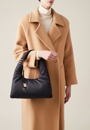 Schwarze Hobo-Tasche mit strukturiertem Finish, runder Form, goldenen Hardware-Akzenten und einem Reißverschlussdetail, gehalten von einer Person in einem beigen Mantel.