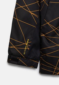 Svart jacktyg med gula abstrakta geometriska mönster. Kardborrestängning vid ärmarna med märket "ICI C'EST PARIS" i gul text.