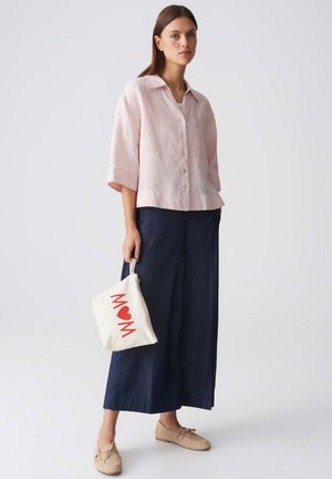 Femme portant une chemise rose pâle à boutons, un pantalon large bleu marine, des mocassins beiges, tenant une pochette blanche avec le texte rouge "MOM" et un symbole de cœur.