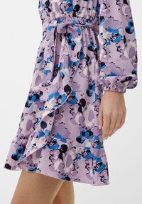 Robe fleurie en lilas avec des accents bleus et noirs. Présente des manches ballons, une ceinture à la taille et un ourlet à volants pour plus de texture.