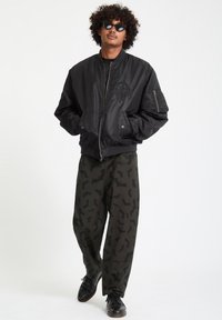 Giacca bomber nera con finitura testurizzata e un design ricamato intricato. Pantaloni verde-grigio con un motivo a pipistrello. Scarpe nere e occhiali da sole.