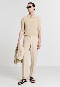 Beige Poloshirt mit strukturiertem Muster, kombiniert mit beigen Nadelstreifenhosen. Das Model hält einen passenden Blazer in der Hand und trägt schwarze Sandalen sowie Sonnenbrille.