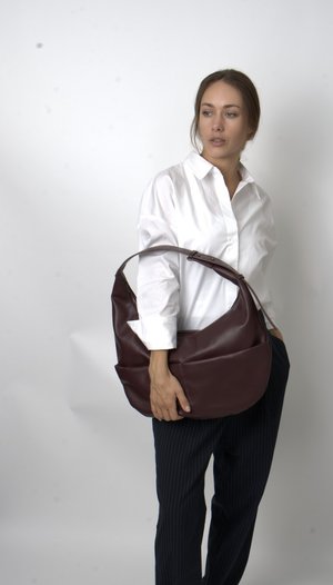 Bolsa de ombro de couro marrom com um design descontraído, apresentando uma alça larga e textura suave. Usada de forma casual com uma camisa branca e calças listradas.