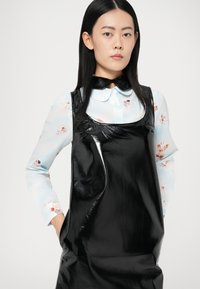 Γυναίκα με ίσια μαύρα μαλλιά φοράει ανοιχτό μπλε μπλούζα με σχέδιο χερουβείμ κάτω από μαύρο λαμπερό αμάνικο φόρεμα, με τα χέρια στις τσέπες.