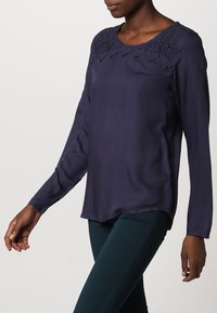 Marinblus med lång ärm och broderad blommönster vid halsringningen, texturerad tyg, avslappnad passform och en lätt asymmetrisk nederkant. Framhävda ärmar.