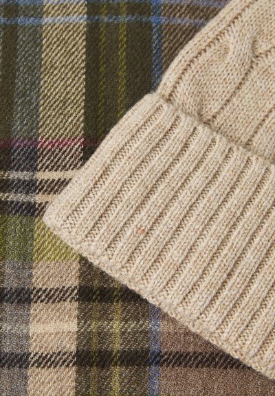 Cappello lavorato a maglia beige con texture a costine, posizionato su un tessuto a quadri con motivi verdi, marroni e blu. Materiali morbidi con texture diverse.