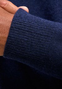 Marineblauer Pullover mit geripptem Muster an den Ärmelbündchen, aus weichem Stoff gefertigt. Nahaufnahme zeigt eine Hand, die auf dem Ärmel ruht, um Details zu verdeutlichen.