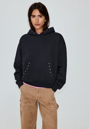 Le modèle porte un sweat à capuche noir à clous par-dessus un haut rose et un pantalon cargo beige. Il se tient devant un fond uni gris clair.