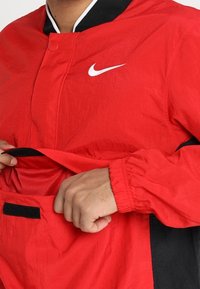 Giacca a vento rossa con accenti neri, dotata di colletto nero, chiusura a scatto e tasca frontale con foderatura in rete. Logo Nike sul petto.