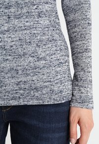 Dlouhý rukáv, pletený top ve námornickém modrém a šedém melírovaném vzoru. Měkká textura s přiléhavým designem, doplněný tmavými džíny.