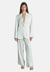 Costume deux pièces blanc en tissu texturé, comprenant un long blazer avec un décolleté plongeant et un pantalon à jambes larges. Des accessoires argentés sont visibles.